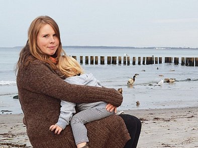 Blonde Frau sitzt mit Kleinkind im Arm am Strand.